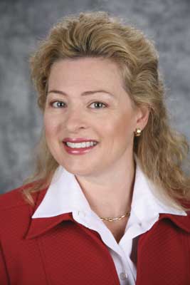 Professional headshot of a woman with curly blonde hair, wearing a red blazer over a white blouse, smiling against a gray background.